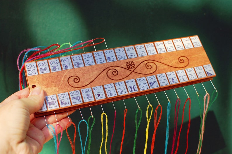 May include: A wooden thread organizer with multiple needles and colorful threads. The organizer has numbers and symbols, with a decorative swirl design. The threads are various colors, including red, blue, and green.