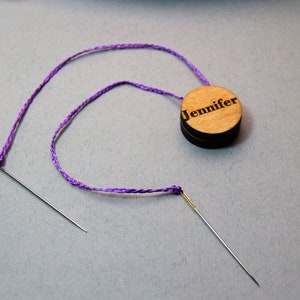 May include: A wooden needle holder with the name "Jennifer" engraved on the top. A purple thread is strung through two needles, one on each end. The needles are silver and the thread is a vibrant purple.