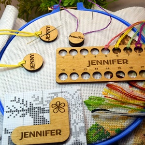 May include: A cross-stitch project in progress, with white fabric stretched in a blue hoop. Wooden tools include needle minders and a thread organizer, all engraved with the name "Jennifer."  Colourful embroidery floss and a pattern are also visible.