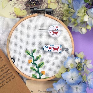 May include: A cross-stitch embroidery hoop with a floral and deer design, along with a needle minder and scissors holder. The embroidery features a green vine with orange flowers. The background includes vintage ephemera and blue hydrangeas.