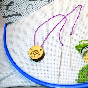 May include: A wooden owl-shaped needle minder with a purple thread attached to two needles. The needles are inserted into a white cross-stitch fabric stretched within a blue embroidery hoop. The design is a craft project.