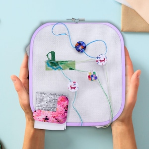 Peut inclure: Un projet de point de croix en cours, maintenu dans un cercle de broderie carré de couleur lavande. Le tissu blanc présente un motif de canard vert partiellement brodé et plusieurs épingles décoratives. Des fils colorés et un morceau de tissu floral sont également visibles.