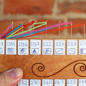 May include: A wooden thread organizer with multiple needles holding colorful threads. The organizer has numbered labels with small drawn symbols. The background is a brick wall.