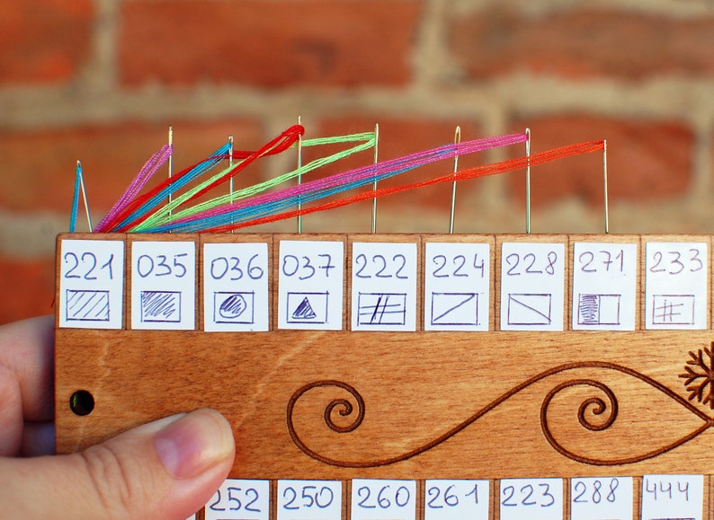 May include: A wooden thread organizer with multiple needles holding colorful threads. The organizer has numbered labels with small drawn symbols. The background is a brick wall.