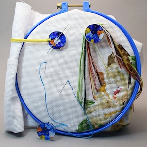 May include: A cross-stitch project in a blue embroidery hoop. The white fabric features a partially completed design with colourful threads. Blue and floral-patterned needle minders hold needles. A white fabric pouch is attached to the hoop.