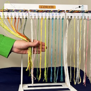 May include: A white wooden thread organiser with multiple coloured threads hanging from it. The organiser has the name "LYNETTE" on it. A hand is holding a bundle of threads in the foreground. The threads are in various colours, including yellow, green, and orange.