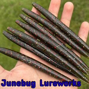 May include: A collection of dark green fishing lures with red and gold speckles, held in a hand against a blurred green background. The lures have a ribbed texture. The text "Junebug Lureworks" is visible at the bottom.