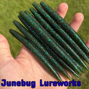 May include: A handful of dark green fishing lures with red and green speckles. The lures are cylindrical and tapered, held against a blurred green background. The text "Junebug Lureworks" is visible at the bottom.