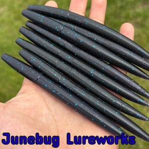 May include: A handful of black fishing lures with teal speckles. The lures are cylindrical with a ribbed texture and a tapered end. The text "Junebug Lureworks" is visible in blue at the bottom of the image.