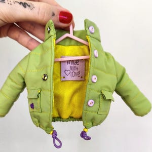 Peut inclure: Une petite veste matelassée verte avec une doublure jaune, des accents violets et un cintre rose. La veste a deux poches et est ornée de boutons violets et d'une étiquette qui dit "Made With Love."