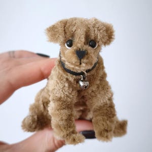 May include: A small, brown plush dog with a black collar and a silver bell. The dog has a fluffy coat, black eyes, and a black nose. It is sitting upright, with its paws in front. The dog is a miniature toy.