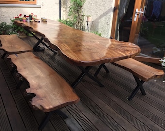 May include: A large wooden table with a live edge top and black metal legs. Two matching wooden benches with live edge tops and black metal legs are placed on either side of the table. The table and benches are on a wooden deck.