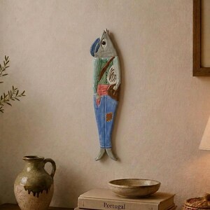 Può includere: Una decorazione da parete verticale in ceramica a forma di pesce con un busto umano. La figura presenta una testa grigia, una giacca verde con una cinghia marrone, una fascia rossa in vita e pantaloni blu con una piccola toppa marrone in basso.