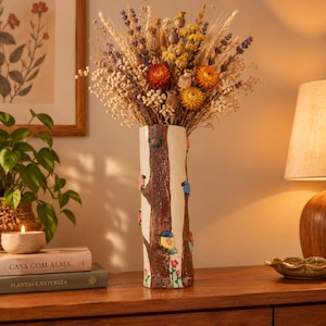 Pode incluir: Um vaso cilíndrico alto de cerâmica com um design de casca de árvore castanha texturizada, apresentando pequenos pássaros e flores coloridas, preenchido com um grande arranjo de flores secas, incluindo lavanda, trigo e flores de palha amarelas e laranjas.