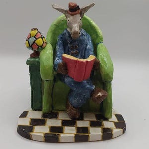 Peut inclure: Une figurine peinte à la main représentant un âne portant une veste bleue et un petit chapeau marron, assis dans un fauteuil vert vif tout en lisant un livre rouge. À côté du fauteuil se trouve une petite lampe à l'abat-jour multicolore.