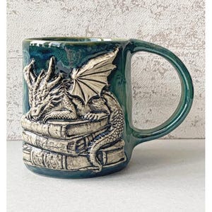 Könnte beinhalten: Eine tealfarbene Keramiktasse mit einem Drachendesign. Der Drache ist in Creme gehalten und sitzt auf einem Stapel Bücher. Die Tasse hat einen großen Henkel und eine glänzende Oberfläche. Ein tolles Geschenk für Fantasy-Fans.