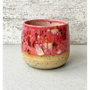 Pode incluir: Caneca de cerâmica artesanal com base bege salpicada e parte superior brilhante. A parte superior apresenta uma mistura de tons vermelhos, rosa e amarelos, criando um design artístico único. A caneca tem uma forma lisa e arredondada.