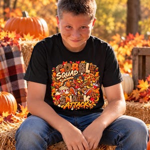 Op de afbeelding: Een zwart t-shirt met korte mouwen en een kleurrijke grafische print van een kalkoen met een zonnebril en een pet, omringd door etenswaren zoals pizza, popcorn en taart, met de tekst "GOBBLE SQUAD SNACK ATTACK" in vetgedrukte letters.
