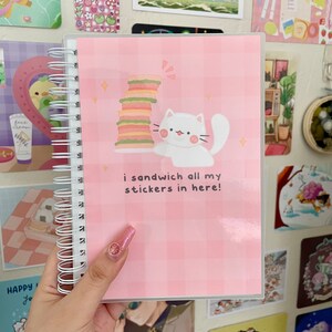 Puede incluir: Un cuaderno con espiral y cubierta rosa a cuadros. La portada presenta un gato de dibujos animados blanco y una pila de sándwiches. El texto "i sandwich all my stickers in here!" está impreso debajo. El cuaderno está en una mano.
