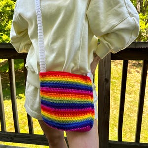 May include: A handmade, rainbow-striped crochet crossbody bag with a white strap. The bag features vibrant horizontal stripes in red, orange, yellow, green, blue, and purple. The bag is worn over the shoulder.