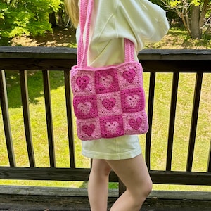 May include: A pink crochet tote bag with a heart pattern. The bag features a square design with nine heart-shaped squares in varying shades of pink. The straps are also pink. The bag is carried over the shoulder.