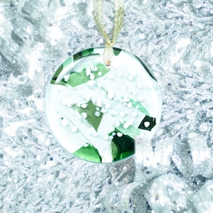 May include: A green and white glass ornament with a white snowflake design. The ornament is hanging from a gold ribbon against a silver background.
