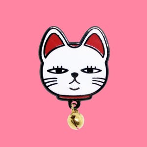 May include: A white cat face pin with red accents on the ears and collar, outlined in black. The cat has closed eyes and whiskers, with a small gold bell dangling below. The pin is set against a pink background.