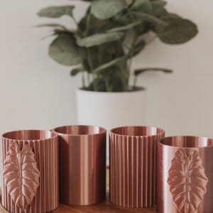 May include: Four cylindrical containers with a metallic copper finish. Two containers feature a vertical ribbed texture, while the other two display a prominent embossed leaf design on the exterior surface. The items are arranged in a row.