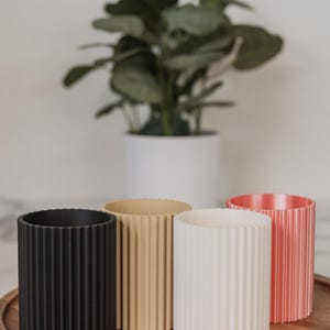 Op de afbeelding: Een set van vier cilindervormige plantenbakken met een verticale geribbelde textuur. De plantenbakken staan op een rij en zijn uitgevoerd in vier verschillende kleuren: matzwart, lichtbeige, gebroken wit en zacht koraalroze.