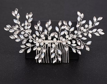 May include: A silver-toned hair comb with a floral design. The comb features clear, oval-shaped crystals and smaller round crystals, arranged to resemble leaves and flowers. The comb's teeth are visible at the base, and the overall design is symmetrical.