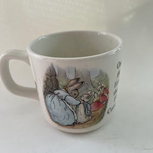 May include: A white ceramic mug featuring a colourful illustration of a rabbit in a blue coat interacting with smaller rabbits in a forest setting. The mug includes printed text that reads, Once upon a time there were four little Rabbits, and their names were Flopsy, Mopsy, Cotton-tail, and Peter.