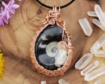 May include: A handcrafted copper wire-wrapped pendant with a black and white fossilized ammonite stone. The pendant hangs on a black cord, surrounded by quartz crystals and flowers. The pendant is a unique piece of jewelry.