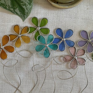 May include: A set of seven stained glass flower suncatchers in various colors, including green, yellow, blue, purple, and pink. The flowers are attached to silver wire stems.
