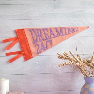 May include: A peach-coloured felt pennant with the words "DREAMING 24/7" in purple lettering. The pennant has an orange border and orange ribbons. The pennant is hanging on a white wooden wall.