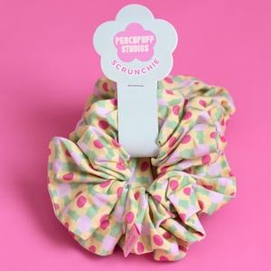 May include: A yellow, green and pink scrunchie with a checkered pattern and pink polka dots. The scrunchie is attached to a white tag with the text "Peachpuff Studios Scrunchie".