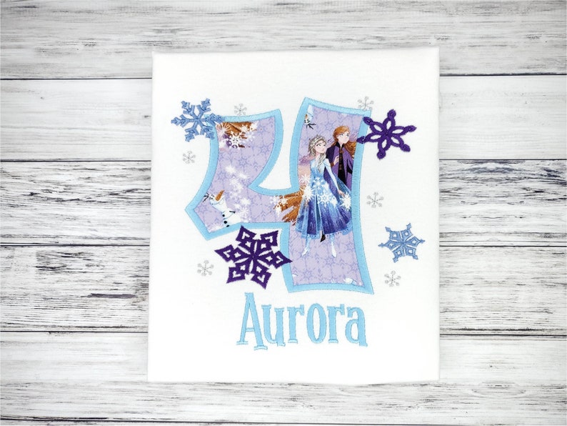 May include: White fabric with a light blue number 4 design, featuring characters from the movie Frozen. The number is outlined in light blue and filled with a snowflake pattern. The name "Aurora" is embroidered in light blue below the number, with snowflake accents.