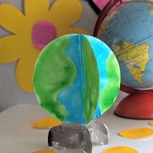 以下が含まれることがあります： 青と緑の水彩画で描かれた手作りの紙地球儀。灰色の台座に取り付けられています。 黄色の花びらと地球儀が背景にあります。 手作りのクラフトプロジェクトです。