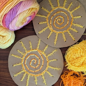 Puede incluir: Dos piezas circulares de cartón marrón con diseños de sol bordados en hilo amarillo y naranja. Bolas de lana amarilla, naranja y multicolor están dispuestas alrededor de los soles, lo que sugiere un proyecto de manualidades o un kit de bricolaje.