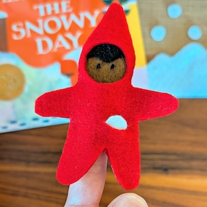 May include: A handmade red felt figure in the shape of a star, with a black face and a white spot. The figure is held up by a finger, with a book titled "The Snowy Day" in the background.