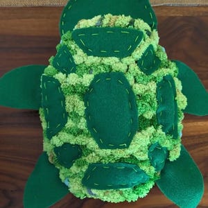 Può includere: Un giocattolo a forma di tartaruga in peluche con un guscio realizzato in pompon di lana verde e lime, arricchito da toppe in feltro verde scuro cucite sulla superficie. La testa e le quattro pinne sono realizzate in feltro verde scuro.