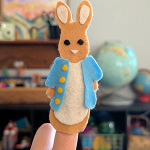 May include: A felt finger puppet of a bunny rabbit, featuring a light brown body, white belly, and long ears with white inner ears. The rabbit wears a light blue jacket with yellow buttons. The puppet is designed to fit on a finger.