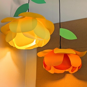 May include: Two hanging pendant lamps shaped like flowers. The left lamp features bright yellow petals with a green leaf on the cord. The right lamp has orange and yellow petals with a green leaf on the cord. Both lamps hang from black cords.