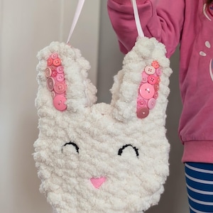Può includere: Una borsa bianca e soffice a forma di coniglio con accenti di bottoni rosa sulle orecchie e un naso a forma di cuore rosa. La borsa ha occhi cuciti neri e un manico a nastro bianco. La borsa è tenuta da una persona.