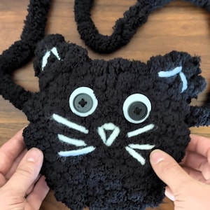 May include: A black cat-shaped bag made of fuzzy, textured yarn. The bag features two white stitched ears, a white stitched nose, and six white stitched whiskers. The eyes are composed of two large, round, light-colored buttons with black centers.
