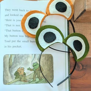 May include: Two frog-eye headbands, one orange and one green, with black pupils and white irises, sit atop an open book. The book's pages display text and a frog illustration. The headbands are made of felt and attached to black headbands.