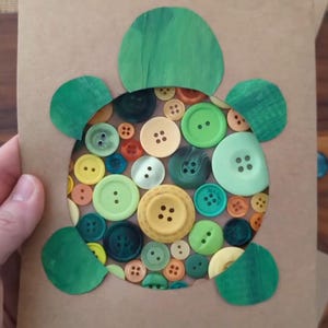 Puede incluir: Una tarjeta rectangular marrón con un recorte circular relleno de un collage de varios botones redondos en tonos verdes, amarillos y naranjas. La tarjeta está decorada con cuatro formas de papel pintadas de verde que representan aletas y una cabeza.
