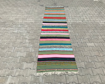 May include: A long, narrow rug with a colorful striped pattern. The rug is made of woven fabric and has a fringe on both ends. The rug is laid out on a gray concrete surface.
