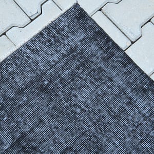 May include: A rectangular area rug featuring a distressed, mottled pattern in shades of black and dark charcoal gray. The rug has a woven texture with visible fibers and a short, straight fringe along the edge, showcasing a uniform, dense weave.