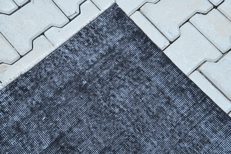 May include: A rectangular area rug featuring a distressed, mottled pattern in shades of black and dark charcoal gray. The rug has a woven texture with visible fibers and a short, straight fringe along the edge, showcasing a uniform, dense weave.