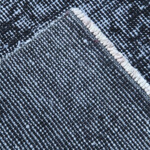 May include: A close-up view of the underside of a rug, showing the dark charcoal gray woven backing material with a distinct grid-like texture and a thick, light cream-colored stitched edge binding that runs along the diagonal corner seam.